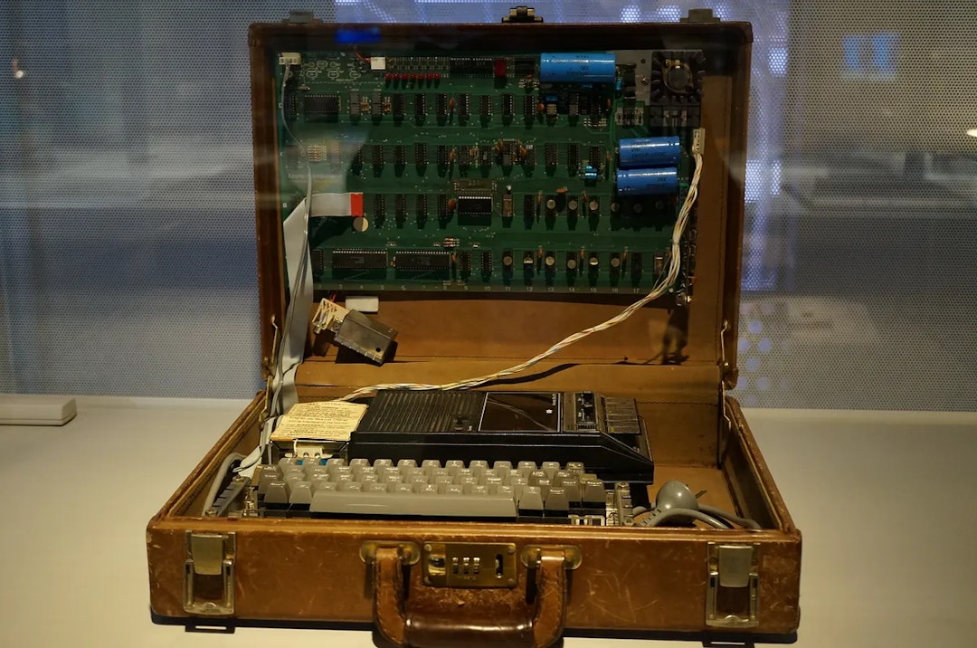 A home built computer with exposed chips and wiring sits in display case in a museum.