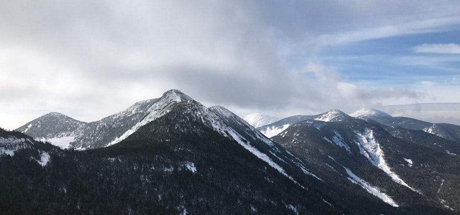 Snowy tree covered mountains stretch off into the distance with dark clouds piling up over top of them.