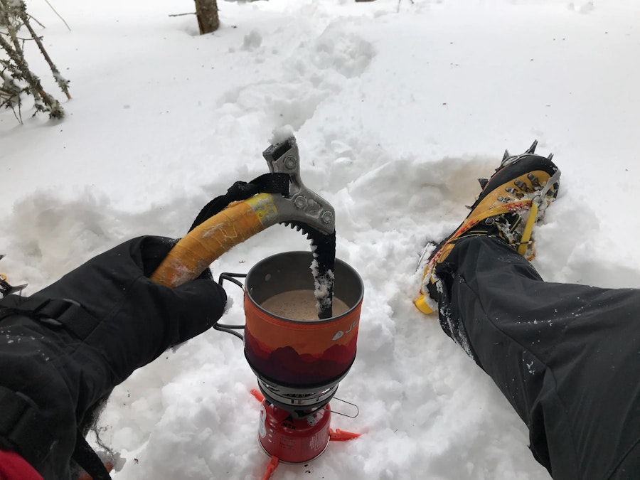 A mountaineer stirring a hot drink over small gas stove with an ice climbing tool.