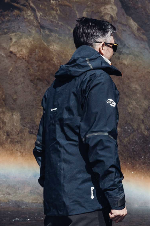 A man wearing a branded rain shell, stands in front of a waterfall.