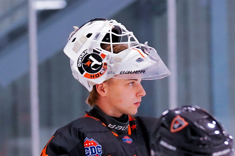 A hockey goalie stands at ease, in full gear, with his helmet perched up to offer a full view of the rink.