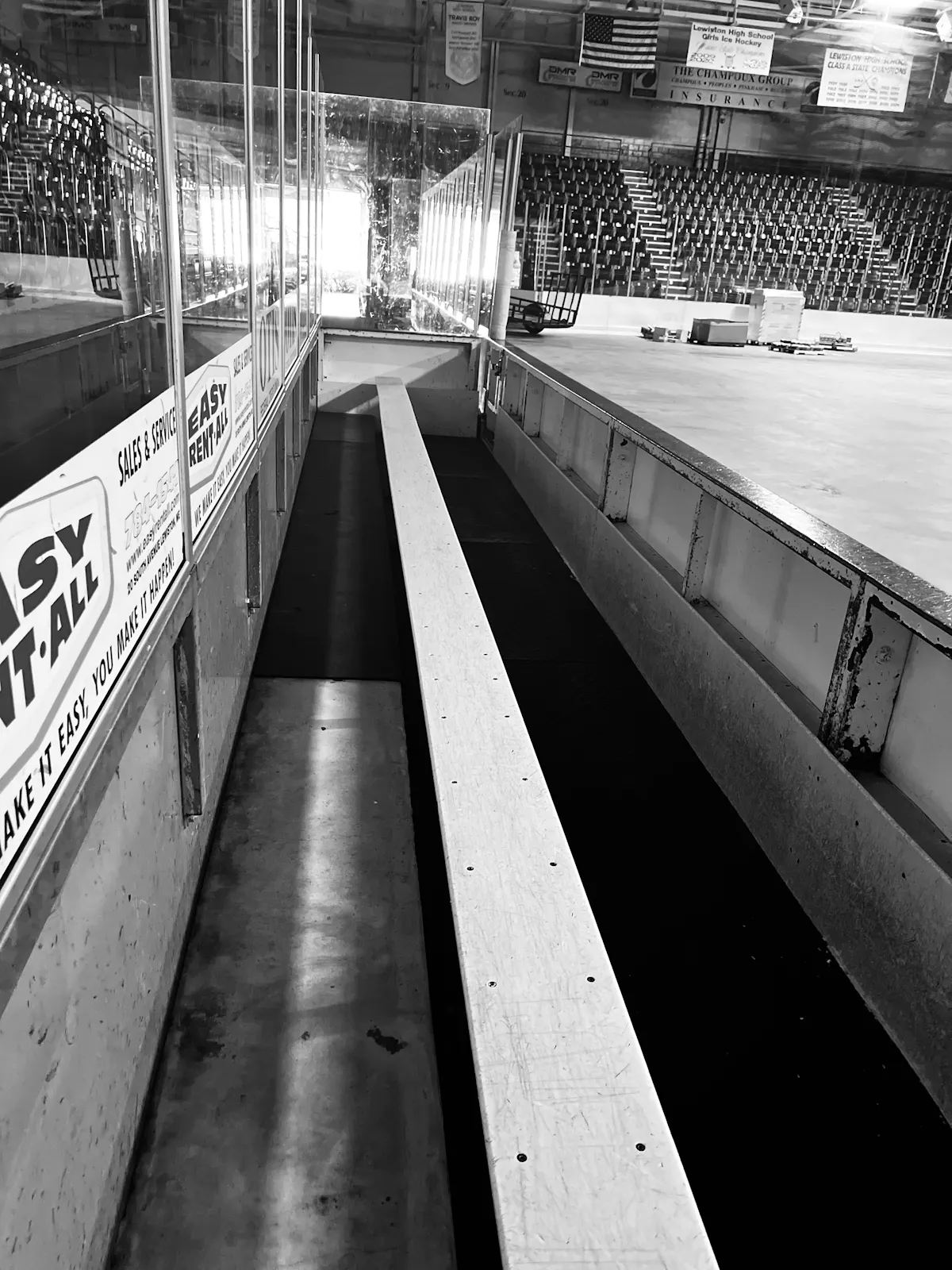 Black-and-white view along an empty wooden hockey team bench beside the ice, enclosed by scuffed boards and plexiglass, with vacant arena seating in the background.