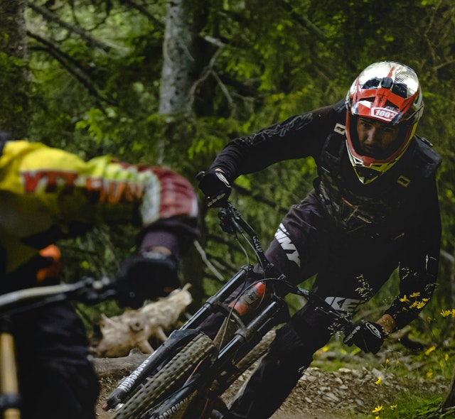 Two mountain bikers wearing full face helmets, caught in motion as they round a corner on a forested track.