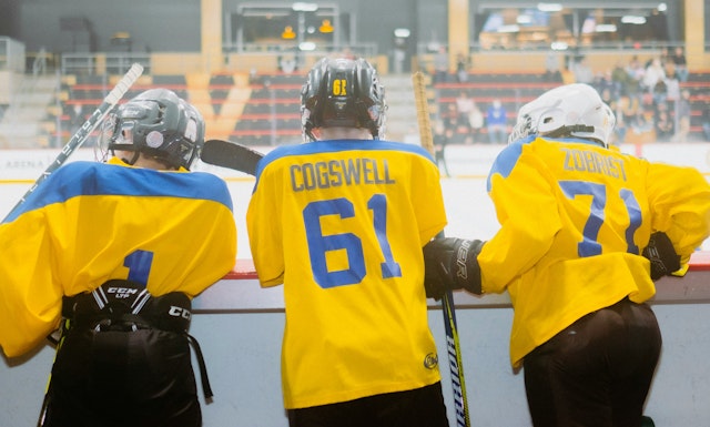 A team of hockey players with their backs to us, facing the ice, waiting for their turn to take the ice.
